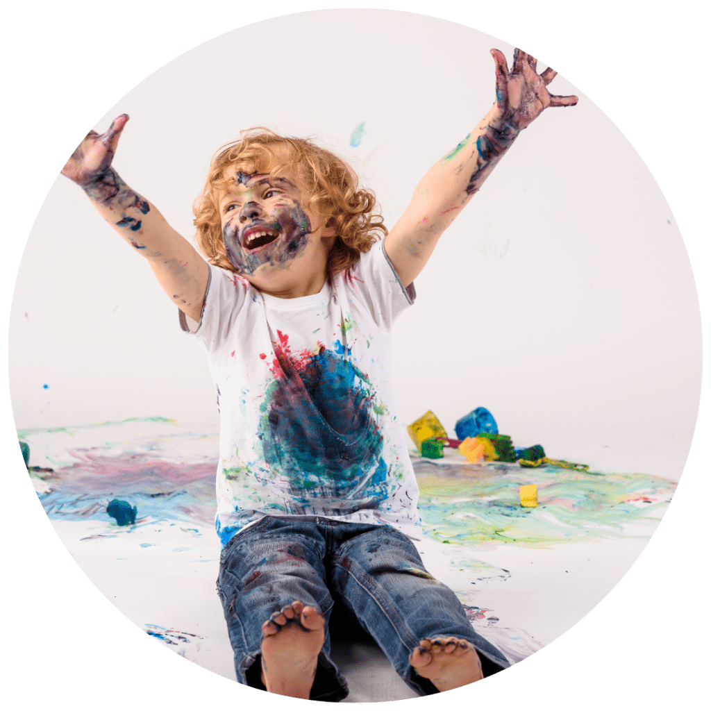 A little boy covered head to toe in paint splotches raises his arms in joy.