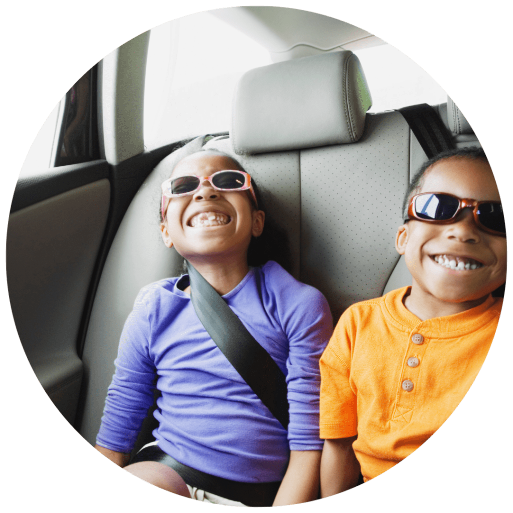 A boy and girl, both wearing sunglasses, smile widely in the backseat of a car.