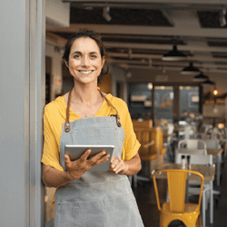 A woman in an apron stands in the doorway of her restaurant with a tablet.