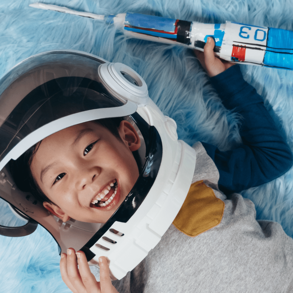 A child wearing a plastic astronaut helmet holds a paper-crafted rocket.