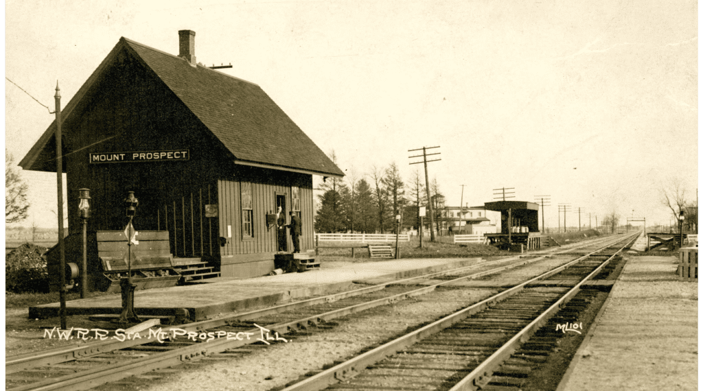 	
Postcard showing Mount Prospect's first railroad depot/station built by Ezra Eggleston in 1873. Sepia print.