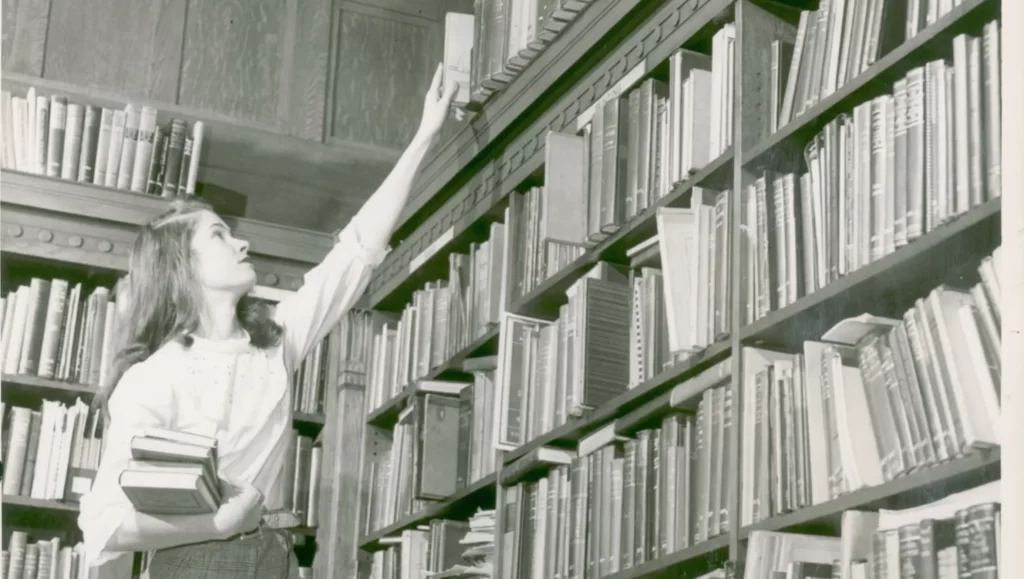 A young woman places a book at the very top of a bookshelf in a library. Black and white.
