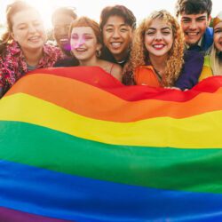 Young diverse people having fun holding an LGBTQ rainbow flag outdoors. Focus on center blond girl.