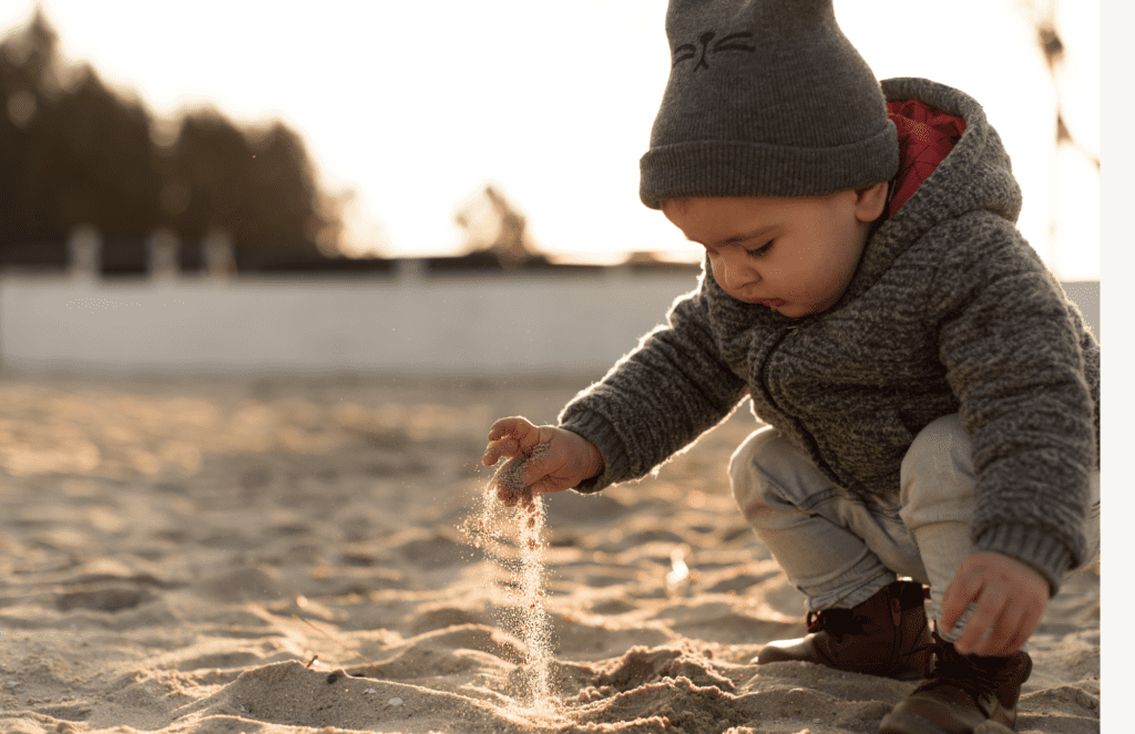 A toddler at the beach in winter watches sand fall from his fingers.