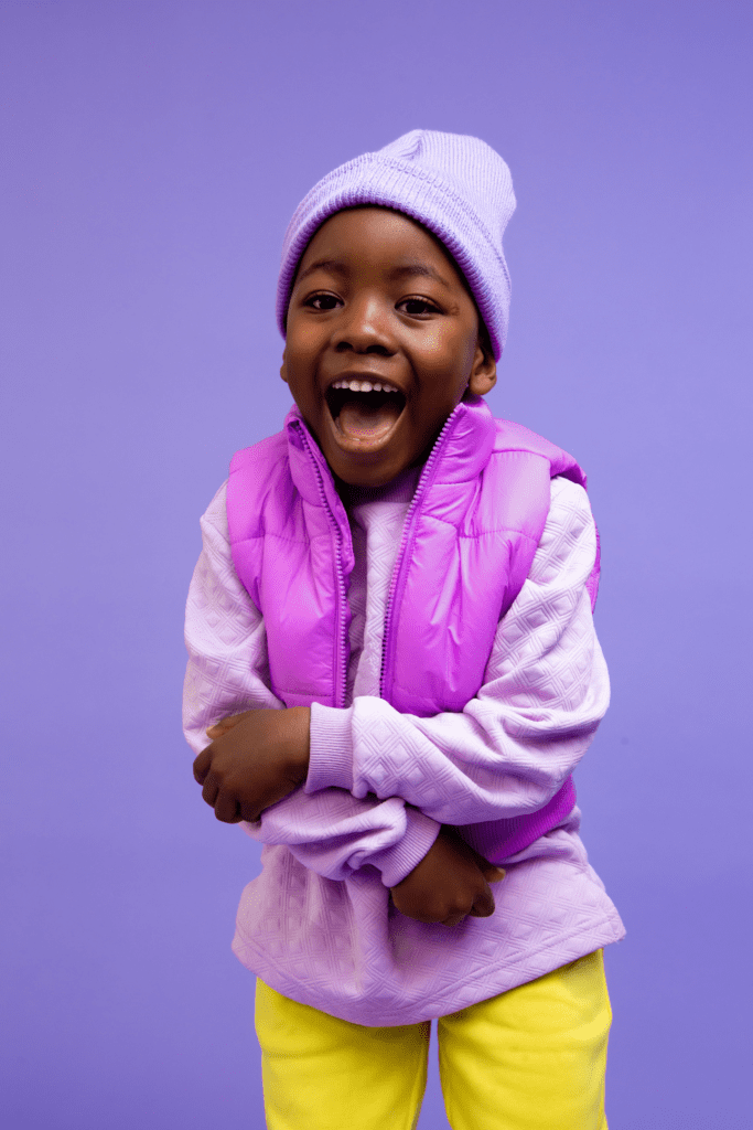 A child with a lavender sweater, bright purple vest, and yellow pants also wears a happy smile.