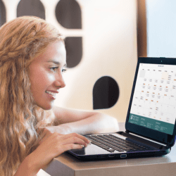 A young woman sits at a table scrolling through a calendar on her laptop.