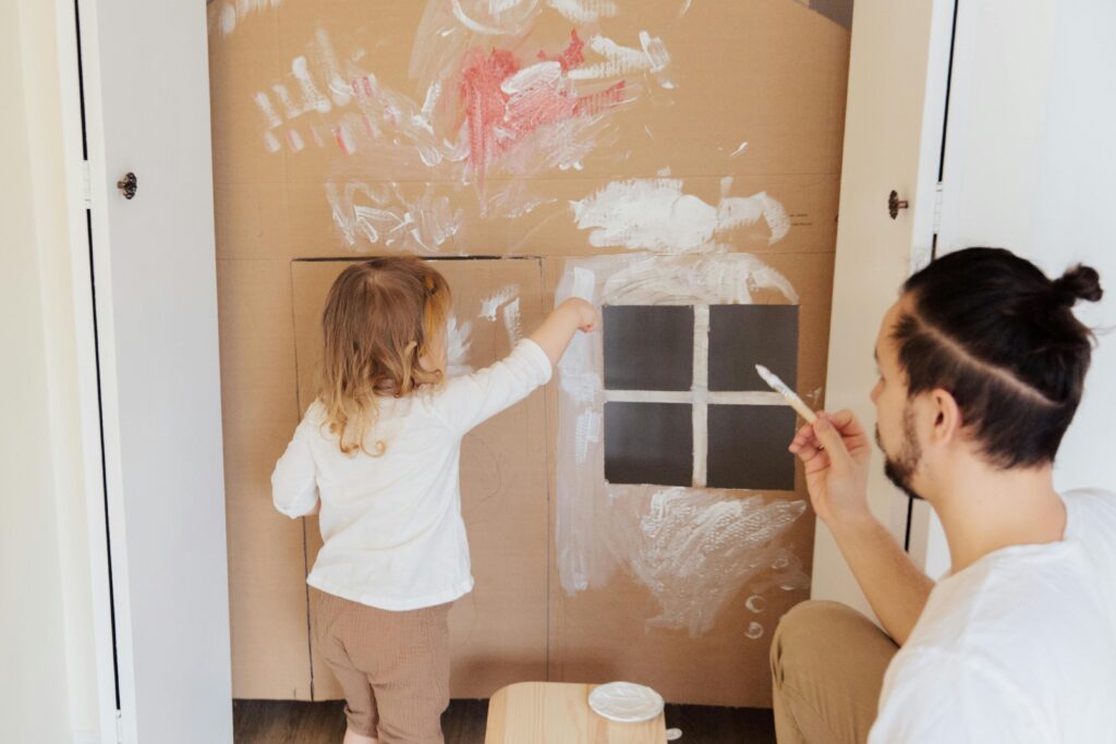 A father and a young child work together to paint a cardboard house that they've made.