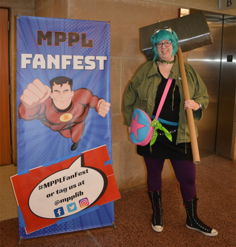 Cosplayer with a turquoise wig and an enormous cardboard hammer stands next to a FanFest sign.